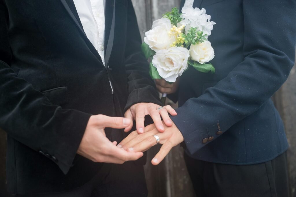 Close-up of two grooms holding hands with wedding rings and bouquet, symbolizing love and commitment.